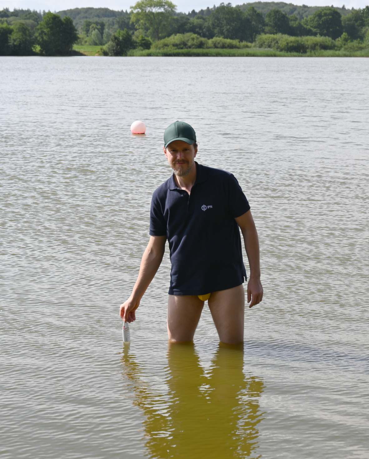 Ein Mann in dunkelblauem Poloshirt, gelber Badehose und grüner Baseballkappe steht bis zu den Knien in einem See und hält ein Thermometer ins Wasser.